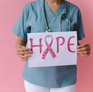 A nurse wearing scrubs with a stethoscope around the neck holds up a sign that reads hope in pink letters. The letter O in hope is a folded pink ribbon